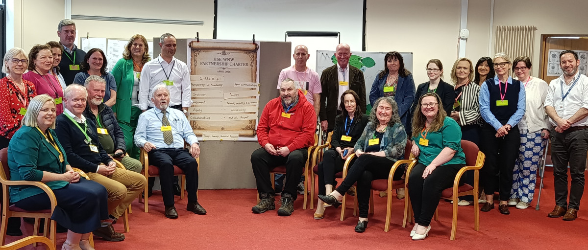 A group of 24 people are in a circle inside a board room, some are sitting some are standing. All are looking at the camera and smling. In between the people is a large board which has a poster attached. The title of the poster is HSE WNW Partnership Charter